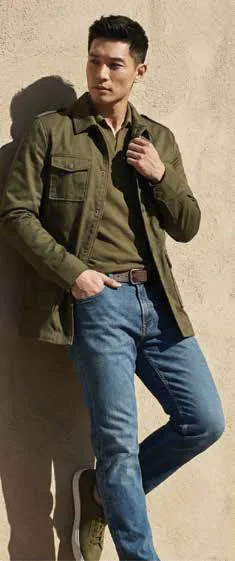A man in an olive shirt-jacket and jeans leaning against a wall