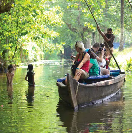 Travellers in a wooden canoe poled along a green backwater, with children standing in the water
