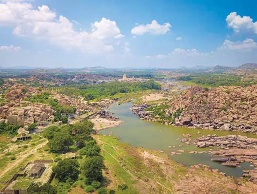 An aerial view of a river winding between boulders and ruins, with a temple tower in the distance