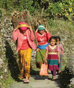Two women carrying bundles of wood on their backs walk a stone village path with two young girls