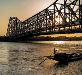 A steel cantilever bridge silhouetted at sunset over a river, with a small boat