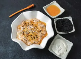 A bowl of seeded porridge with small bowls of honey, black seeds and a white cream