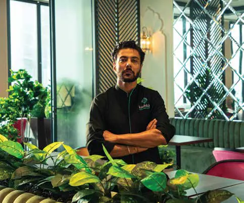 Ranveer Brar in a black chef's jacket, arms crossed, seated behind leafy plants in a restaurant