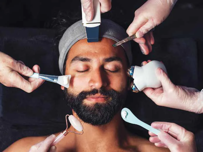 A bearded man receiving a facial treatment from several hands holding tools