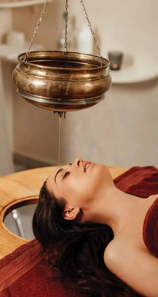 A woman receiving a Shirodhara treatment, oil pouring from a brass vessel onto her forehead
