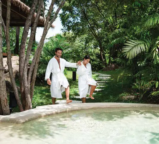 A couple in white robes walking beside an outdoor pool among trees