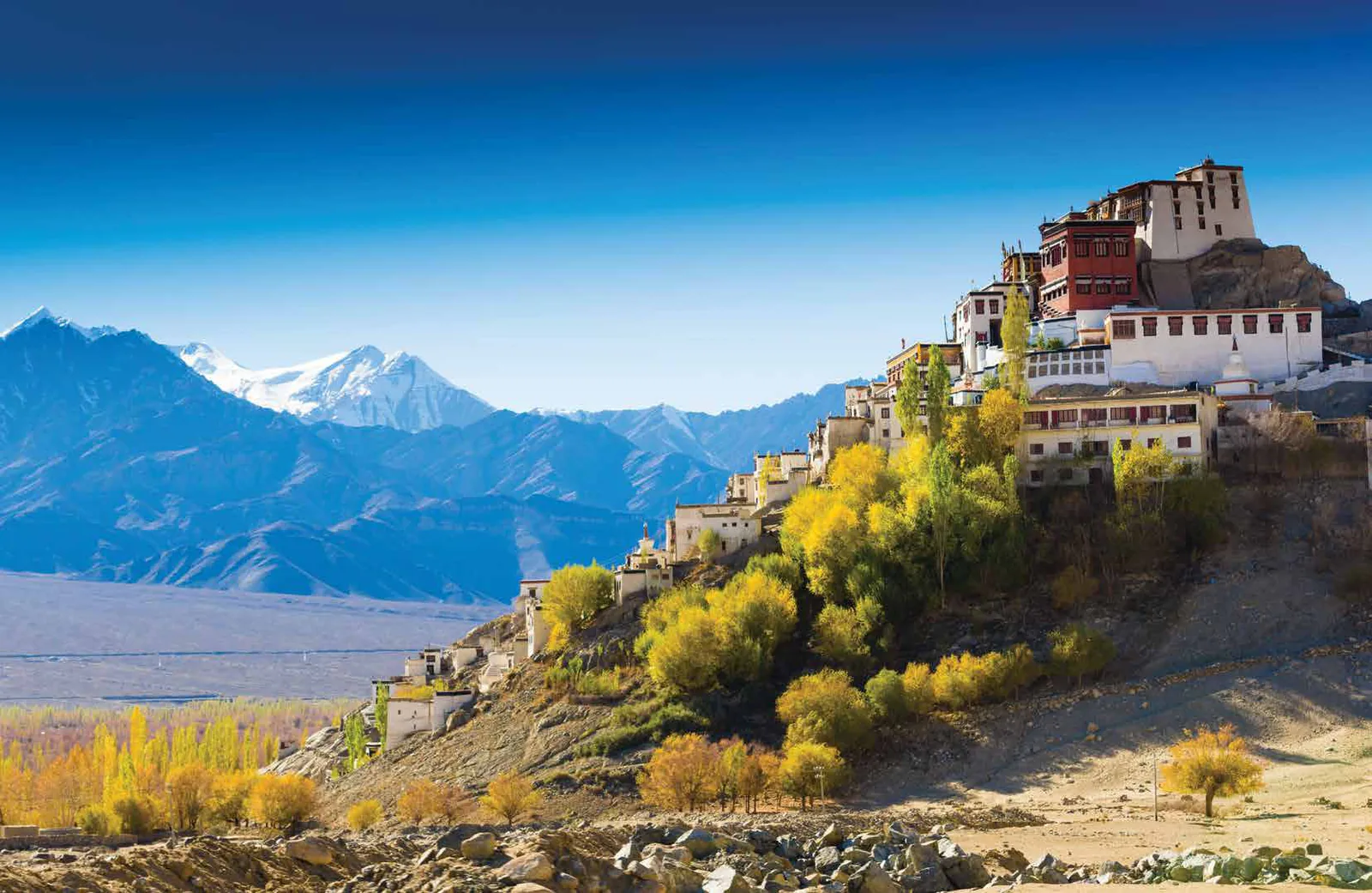 A whitewashed hilltop monastery above autumn trees, with snow-capped mountains behind