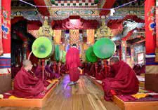 Monks in maroon robes seated in a monastery hall hung with large drums