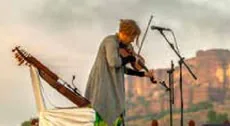 A musician playing the violin on an open-air stage, with a fort behind