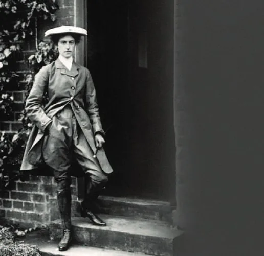 A vintage photograph of a woman in a hat, long coat and boots standing in a doorway