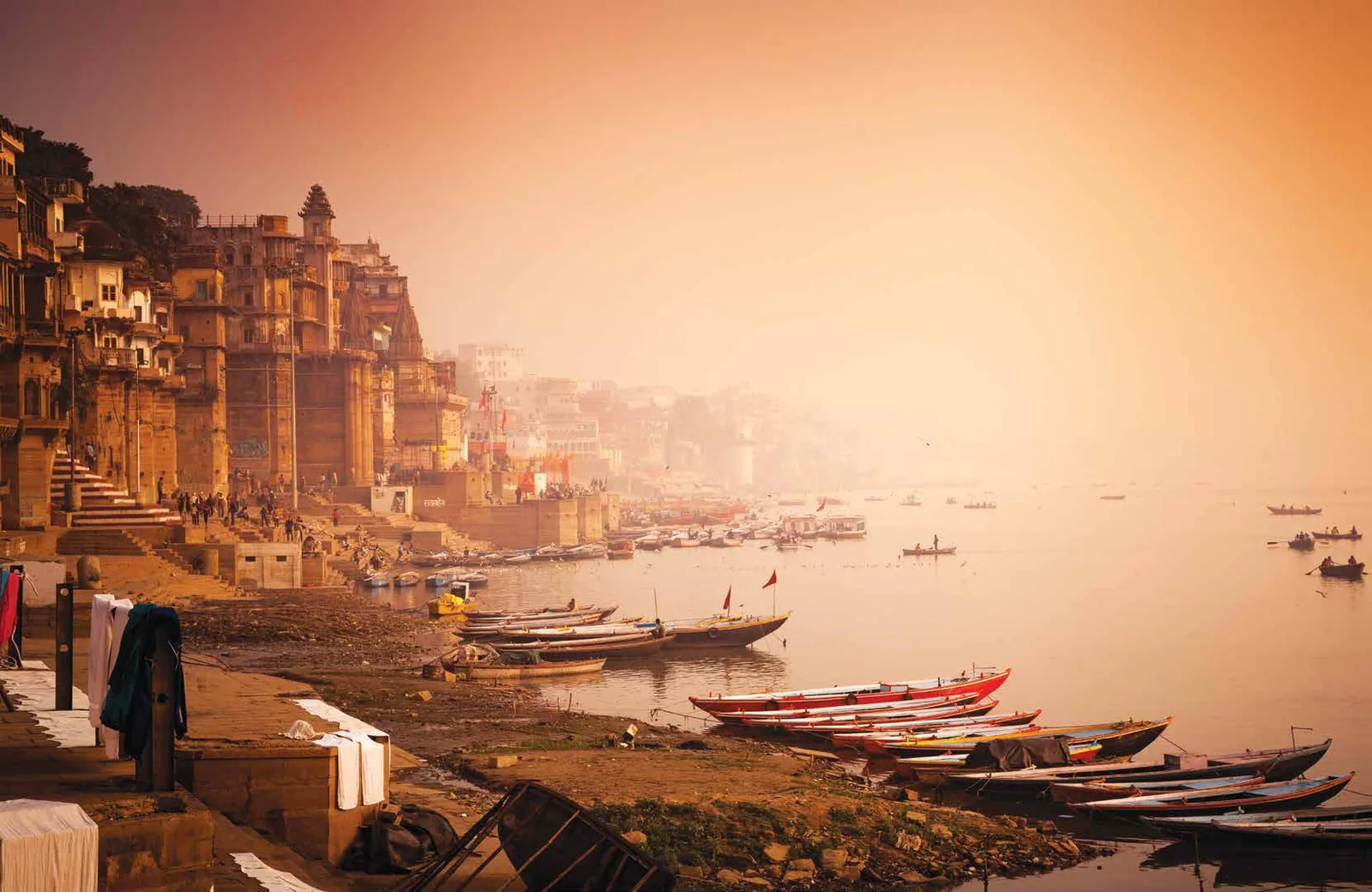 Old riverside buildings and ghats in hazy golden light, with boats moored along the river