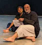 Two actors seated on the floor of a stage